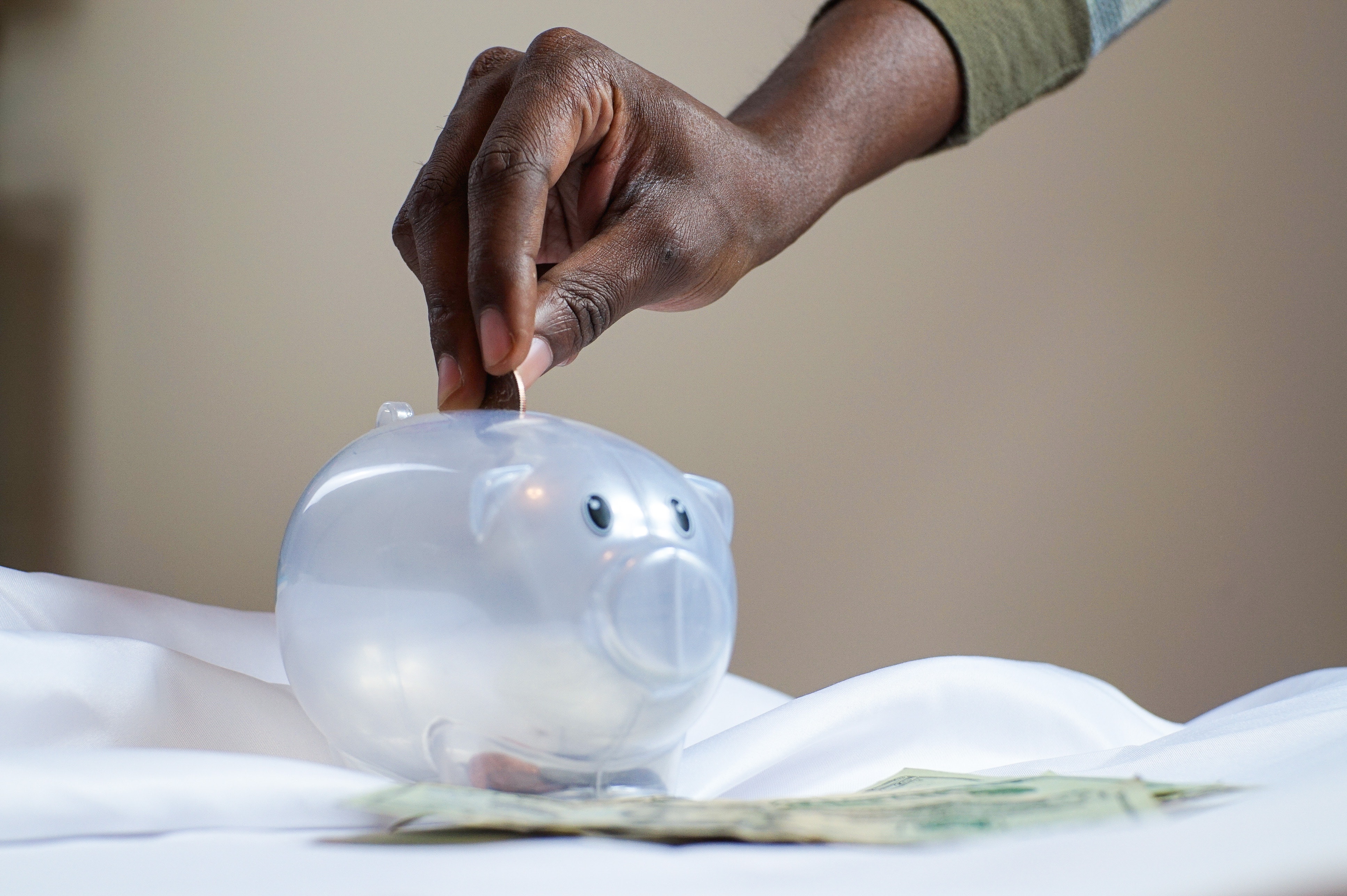 hand adding coin to clear piggy bank sitting on a white table cloth.